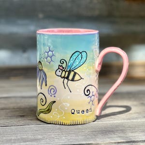 May include: A ceramic mug with a pink handle and rim, featuring a hand-painted design. The design includes a bee with a crown, flowers, and the word "Queen". The mug has a light blue and yellow background.