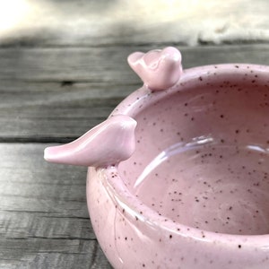 May include: A pink ceramic bowl with a speckled finish and two small bird-shaped handles. The bowl is perfect for holding small items like jewelry or trinkets.