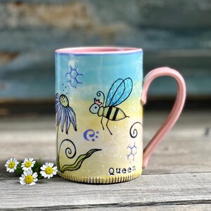 May include: A ceramic mug with a light blue and yellow gradient background featuring a hand-painted bee wearing a crown, a purple flower, and honeycomb patterns. The word Queen is printed near the base. The mug has a pink handle and interior.