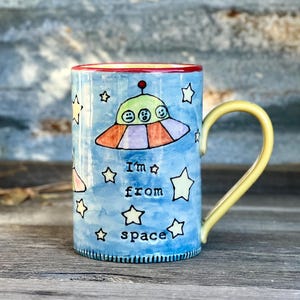 May include: A ceramic mug with a blue background and a yellow handle. The mug features a colorful illustration of a UFO with three alien faces and the text "I'm from space" surrounded by stars. The rim is red.