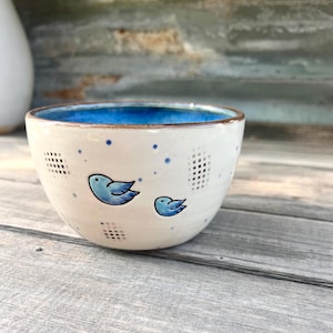 May include: A ceramic bowl with a white exterior, featuring two blue bird illustrations and blue dots. The interior of the bowl is a vibrant blue. The bowl has a brown rim and is set on a wooden surface.