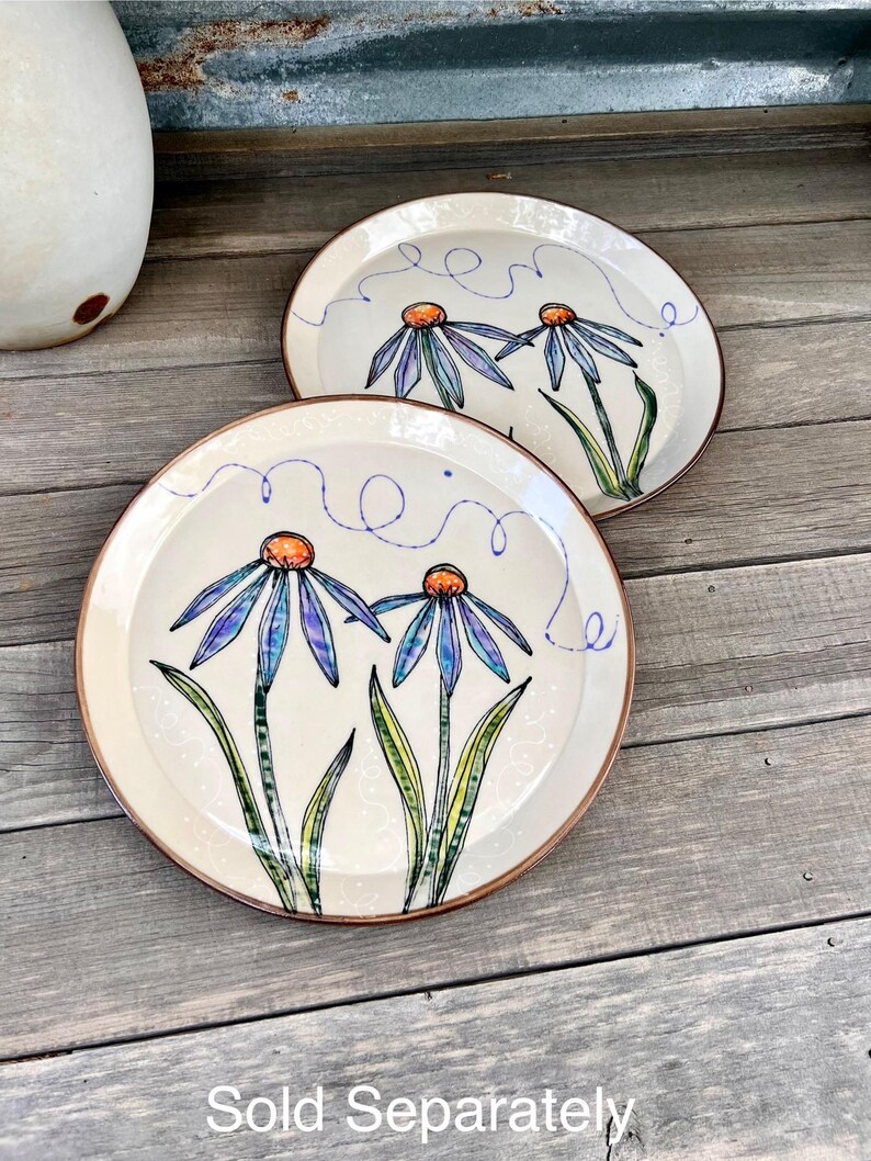 May include: Two cream-colored ceramic plates with a hand-painted floral design. Each plate features blue and purple flowers with orange centers and green stems. The plates have a brown rim and are on a wooden surface. The text "Sold Separately" is at the bottom.