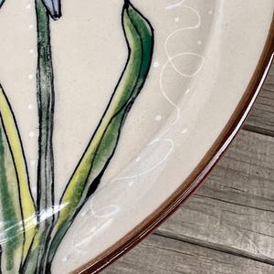 May include: A ceramic plate with a floral design. The plate features a cream-colored surface with a brown rim. The floral design includes blue and green flowers with green leaves. White dots and swirls add detail to the design.