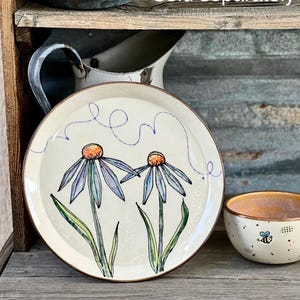 May include: A cream-colored ceramic plate with a brown rim, featuring two stylized blue and purple flowers with orange centers. A matching bowl with a bee design is also visible. The words "Sold separately" are in the image.