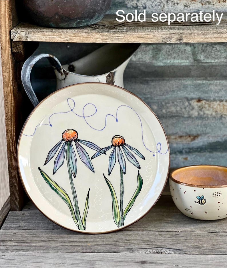 May include: A cream-colored ceramic plate with a brown rim, featuring two stylized blue and purple flowers with orange centers. A matching bowl with a bee design is also visible. The words "Sold separately" are in the image.