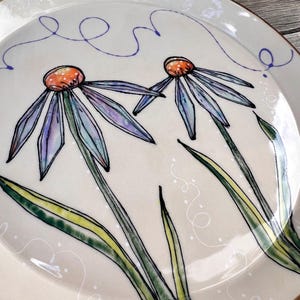 May include: A cream-colored ceramic plate with a hand-painted floral design. Two large flowers with purple and blue petals and orange centers are depicted. Green leaves and stems are also visible. The plate has a brown rim and a whimsical blue line design.