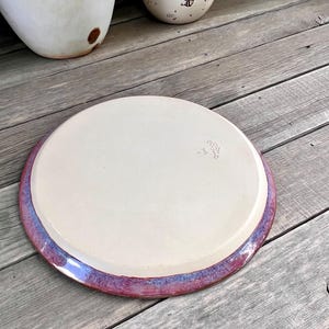May include: A handmade ceramic plate with a cream-colored center and a vibrant purple and blue rim. The plate is round and appears to be sitting on a wooden surface. The plate has a unique, artistic design.
