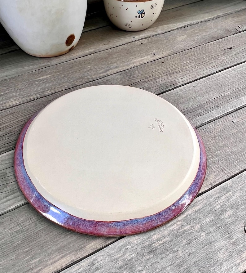 May include: A handmade ceramic plate with a cream-colored center and a vibrant purple and blue rim. The plate is round and appears to be sitting on a wooden surface. The plate has a unique, artistic design.