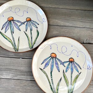May include: Two off-white ceramic plates with hand-painted blue and purple flowers with orange centers and green leaves. Each plate has a brown rim and a blue swirl design. The plates are on a wooden surface.