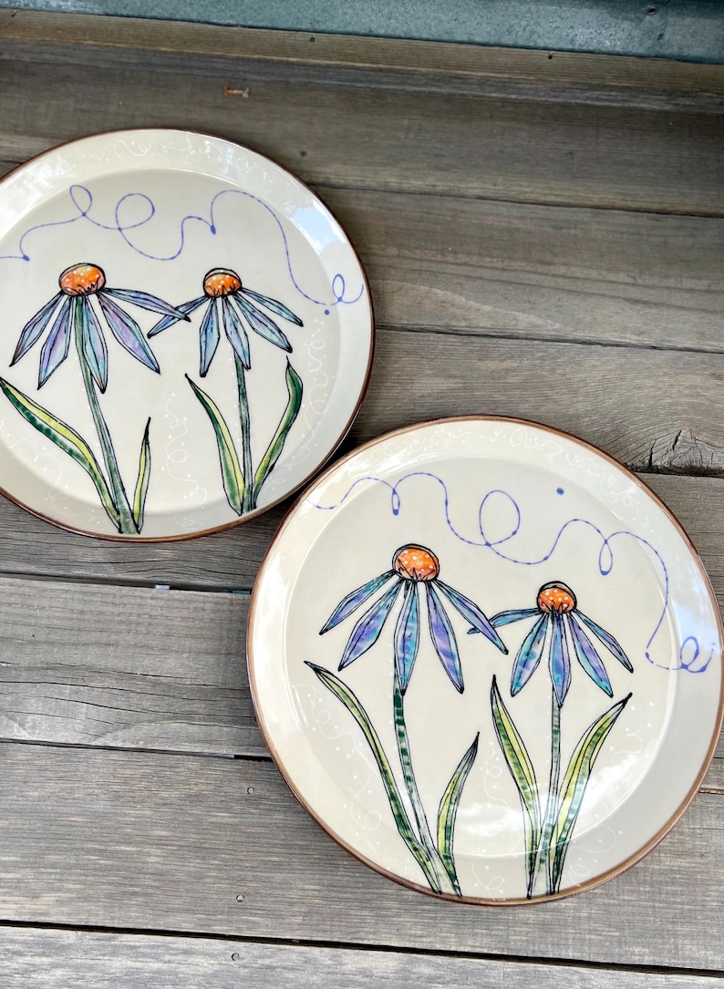 May include: Two off-white ceramic plates with hand-painted blue and purple flowers with orange centers and green leaves. Each plate has a brown rim and a blue swirl design. The plates are on a wooden surface.