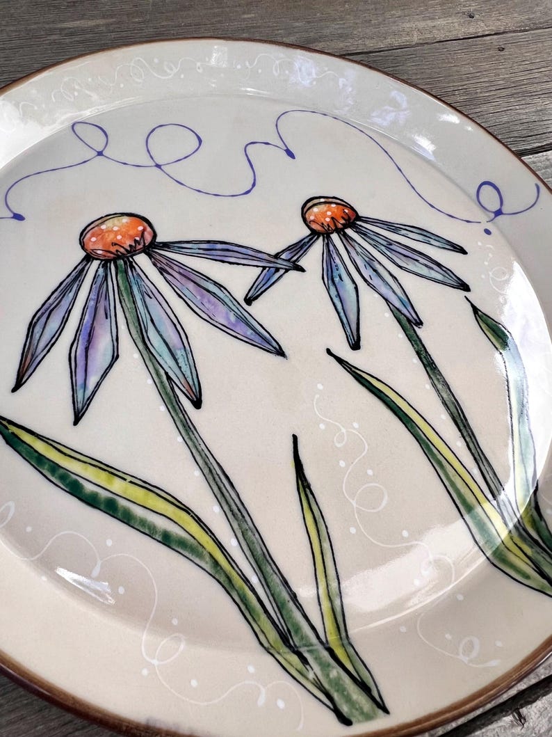 May include: A cream-colored ceramic plate with a hand-painted floral design. Two large flowers with purple and blue petals and orange centers are depicted. Green leaves and stems are also visible. The plate has a brown rim and a whimsical blue line design.