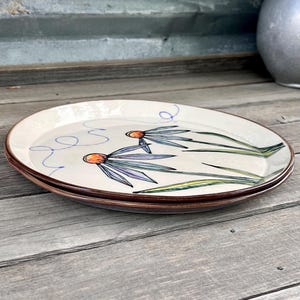 May include: Two oval ceramic plates with a floral design. The plates have a cream-colored background with a brown rim. The floral design features orange centers, blue petals, and green stems. The plates are stacked on a wooden surface.