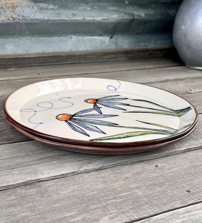 May include: Two oval ceramic plates with a floral design. The plates have a cream-colored background with a brown rim. The floral design features orange centers, blue petals, and green stems. The plates are stacked on a wooden surface.