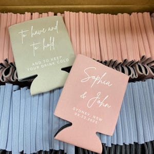 May include: A box of pink and blue can coolers with the text "to have and to hold" and "AND TO KEEP YOUR DRINK COLD" on one cooler and "Sophia & John" and "SYDNEY, NSW 09.23.2025" on another cooler.