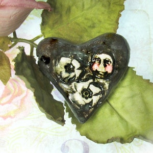 Puede incluir: Un colgante de cerámica en forma de corazón con un diseño floral y dos caras pintadas en él. El colgante es marrón con detalles blancos y rosados y está apoyado en una hoja verde.