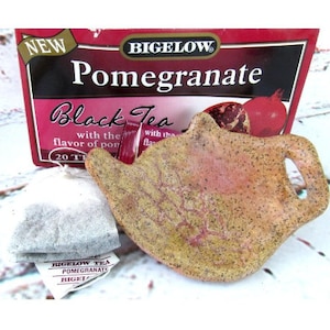 Puede incluir: Una caja de té negro de granada Bigelow con 20 bolsitas de té. La caja es roja con texto blanco. Un infusor de té de cerámica con forma de tetera está en primer plano.