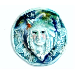 May include: A round ceramic tile with a blue and white glaze. The tile features a raised image of a Native American man's face with a feathered headdress.