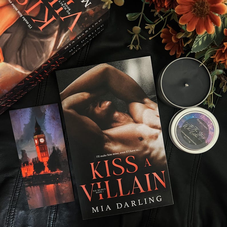 May include: A paperback book titled "Kiss a Villain" by Mia Darling. The book cover features a shirtless man with dark hair and a muscular build. The book is part of a series called "No Mercy" and is book number one. The book is lying on a black surface with a candle and a bookmark.