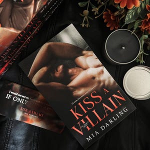 May include: A paperback book titled "Kiss a Villain" by Mia Darling. The cover features a shirtless man lying on a bed with his head turned to the side. The book is on a black leather surface with a silver candle holder and a black candle.