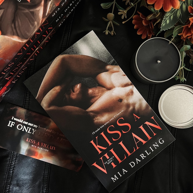 May include: A paperback book titled "Kiss a Villain" by Mia Darling. The cover features a shirtless man lying on a bed with his head turned to the side. The book is on a black leather surface with a silver candle holder and a black candle.