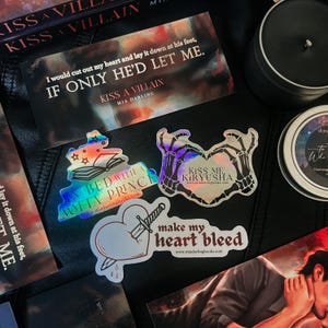May include: A set of five iridescent holographic stickers featuring quotes and illustrations from the book "Kiss a Villain" by Mia Darling. The stickers feature the text "Kiss a Villain", "In Bed with a Mafia Prince", "Kiss Me Kiryusha", "Make My Heart Bleed", and "I would cut out my heart and lay it down at his feet, If Only He'd Let Me."
