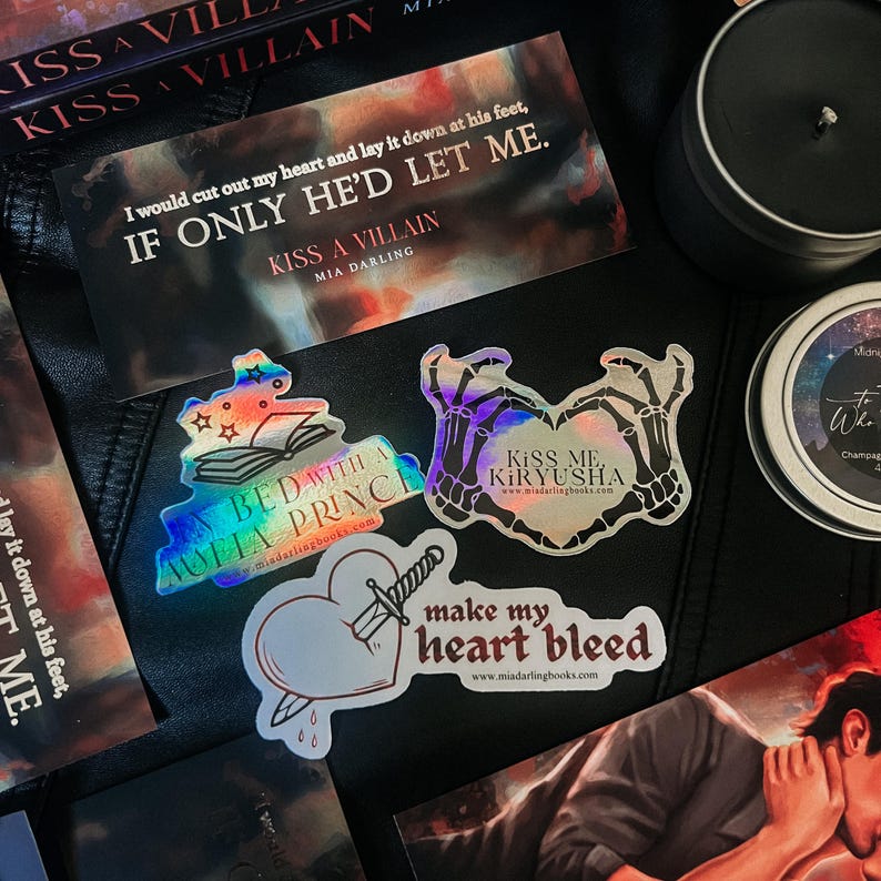 May include: A set of five iridescent holographic stickers featuring quotes and illustrations from the book "Kiss a Villain" by Mia Darling. The stickers feature the text "Kiss a Villain", "In Bed with a Mafia Prince", "Kiss Me Kiryusha", "Make My Heart Bleed", and "I would cut out my heart and lay it down at his feet, If Only He'd Let Me."