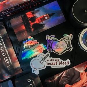 May include: A set of four holographic stickers featuring book-themed designs. The stickers feature a book with stars, a heart with a dagger, a pair of skeletal hands forming a heart shape, and a book with the text "Kiss Me Kiryusha".