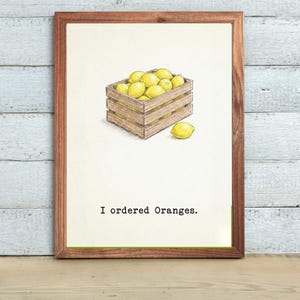 May include: A framed print featuring a wooden crate filled with yellow lemons. One lemon sits outside the crate. The text "I ordered Oranges" is printed below the crate.