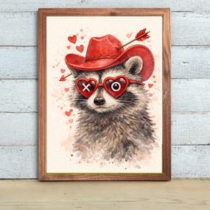May include: Watercolor print of a raccoon wearing a red cowboy hat and heart-shaped glasses with "X" and "O" lenses. An arrow pierces the hat. The raccoon is surrounded by small red hearts, creating a Valentine's Day theme.
