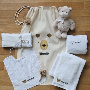 Könnte beinhalten: Ein Baby-Geschenkset mit einem beigen Kordelzugbeutel mit Bärengesicht und dem Namen "Nicolò", einem Teddybären, einem Lätzchen und zwei weißen Handtüchern. Die Artikel haben gestickte Bärendetails und sind auf einer Holzoberfläche angeordnet.