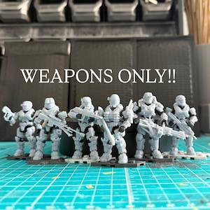 May include: A collection of grey action figures, each armed with a weapon, are positioned on a blue and green surface. The text "WEAPONS ONLY!!" is displayed above. The figures are in a line, with detailed armour.