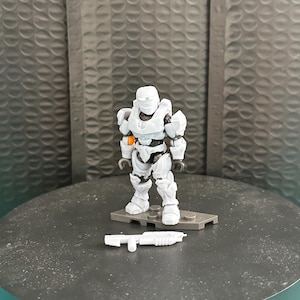 Könnte beinhalten: Eine weiße Plastik-Actionfigur eines Soldaten, der einen Helm und eine Rüstung trägt und eine weiße Plastikwaffe hält. Die Figur steht auf einem grauen Plastiksockel.