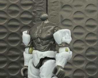 May include: A gray and white action figure of a muscular humanoid robot. The figure has a detailed, textured torso and is wearing white armor on its arms and legs. The figure is posed against a textured gray background.