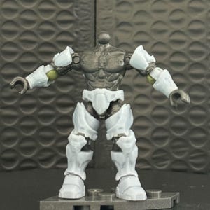 May include: A headless action figure with a gray torso and white armor on the arms, legs, and torso. The figure is standing on a gray base. The figure's arms are bent at the elbows, and the hands are open.