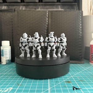 May include: A group of six gray and white miniature figures, possibly soldiers or space troopers, are standing on a black rotating platform. They are detailed with black accents and stand on a green grid surface. The background includes storage containers and art supplies.