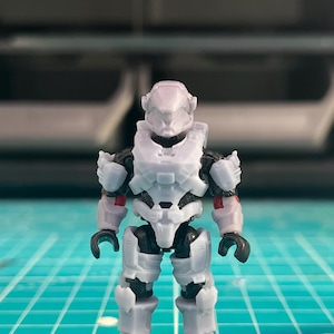 May include: A white and black action figure of a futuristic soldier. The figure has a detailed armor design with red accents on the arms. It stands on a black base against a blurred background.