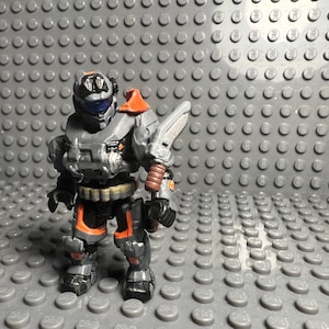 May include: A gray and orange action figure of a soldier wearing a helmet and holding a weapon. The figure is standing on a gray background.
