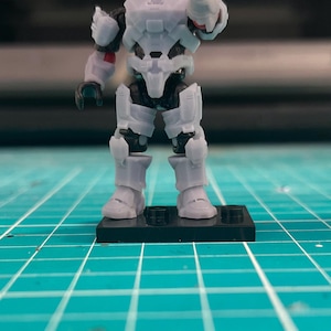 May include: A white and gray action figure of a soldier wearing armor. The figure is standing on a black base with two black studs.