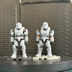 Könnte beinhalten: Zwei graue Actionfiguren von gepanzerten Soldaten, die auf grauen und weißen Plattformen stehen. Die Figuren haben detaillierte Rüstungen und Helme mit einigen orangefarbenen Akzenten. Der Hintergrund ist eine strukturierte, dunkelgraue Oberfläche.