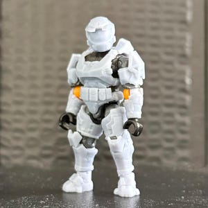 Op de afbeelding: Een lichtgrijs actiefiguur van een futuristische soldaat in volledige bepantsering. Het figuur heeft zwarte en oranje accenten, met een helm en beweegbare gewrichten. Het figuur staat op een donkere ondergrond.