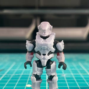 May include: A white plastic action figure of a soldier wearing a helmet and armor. The figure has red accents on the arms and is standing on a black base.