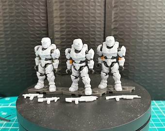 May include: Three gray and white action figures with orange accents stand on a black circular base. Each figure is holding a gray weapon. The figures are on a green surface.