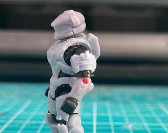May include: A gray action figure of a futuristic soldier, with a helmet and armor. The figure has black accents and a red dot on the arm. It is mounted on a black base, set against a blue and green surface.
