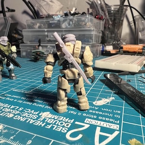 May include: A beige and gray plastic action figure of a soldier holding a sword. The figure is standing on a blue cutting mat with a ruler and the text "CAUTION" printed on it.
