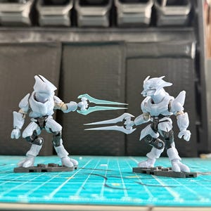 May include: Two light grey action figures, each holding a translucent blue energy sword. The figures stand on black bases, against a blurred background. The figures have black detailing.