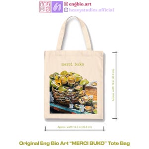 May include: Cream-colored tote bag with the text "merci buko" and a detailed illustration of a basket filled with green fruit. The bag is approximately 16 inches (40.6 cm) high and 14.5 inches (36.8 cm) wide. The bag is labeled "Original Eng Bio Art MERCI BUKO Tote Bag".