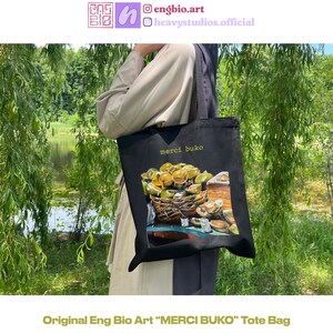 May include: Black tote bag featuring a colorful illustration of a basket of fruit with the text "merci buko". The bag is held in front of a natural background with trees and water. The text "Original Eng Bio Art" is at the bottom.