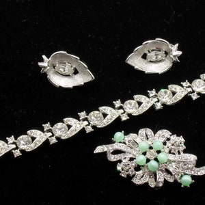 May include: A vintage jewelry set featuring a bracelet, earrings, and a brooch. The silver-toned pieces are adorned with clear rhinestones and light green beads. The earrings are leaf-shaped.