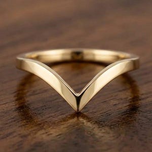 Op de afbeelding: Een gepolijste gouden ring met een V-vormig chevronontwerp. De band heeft een glad, reflecterend oppervlak en een puntig midden, wat zorgt voor een opvallend hoekig silhouet. Het metaal heeft een warme, gele tint met een glanzende afwerking.