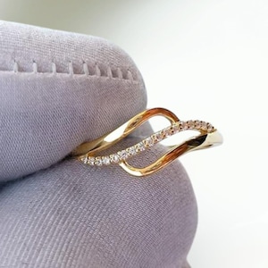 Op de afbeelding: Een gouden ring met een bypass-ontwerp met twee overlappende, golvende banden. De ene band is van gepolijst massief goud, terwijl de andere is bezet met een rij kleine, heldere, sprankelende steentjes die de welving van de structuur volgen.
